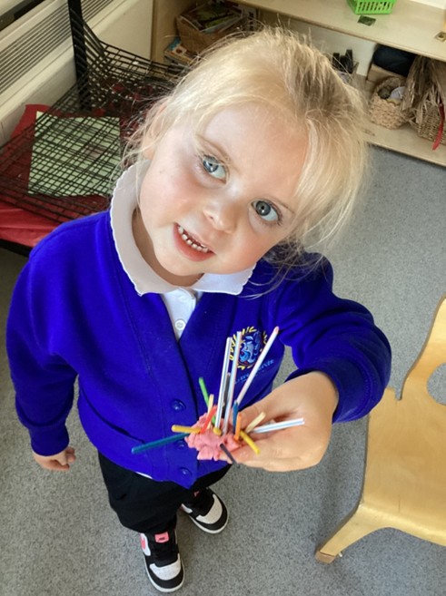 A young girl with blonde hair looks up at the camera while proudly holding out her playdough creation, which is filled with many upright, colorful sticks.