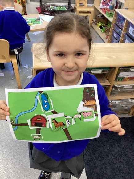 A young girl in a blue cardigan smiles as she displays her completed color farm map, featuring various animals and a red barn glued onto a green landscape.