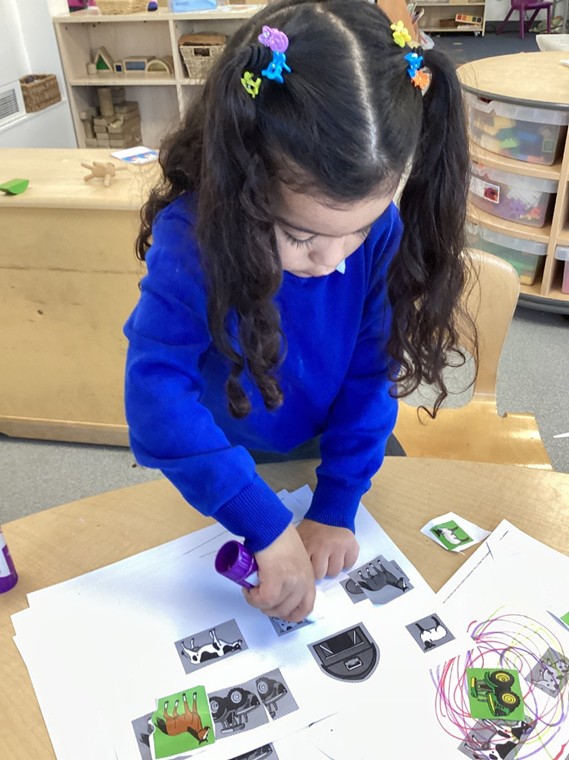 A young girl with long dark hair in pigtails focuses intently on using a purple glue stick to attach farm animal cut-outs to a sheet of paper.
