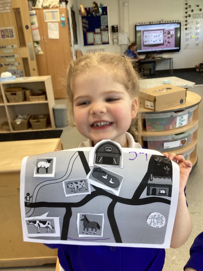 A smiling girl proudly holds up her finished farm map, which features black-and-white icons of a barn, sheep, horse, and pond glued onto a path layout.