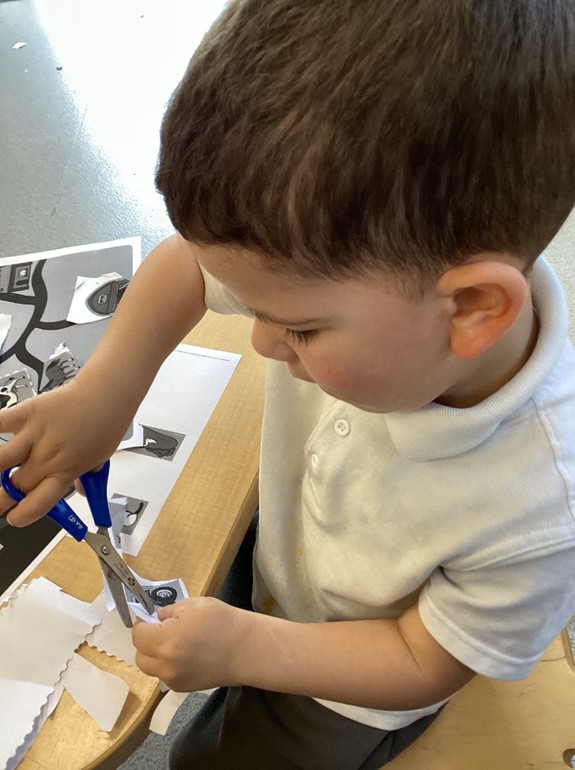 A close-up of a young boy using blue safety scissors to carefully cut out small paper squares featuring farm-related images.