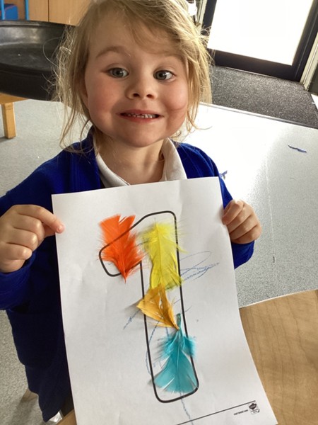 A smiling young girl holds up a large printed number "1" that she has decorated with bright orange, yellow, and turquoise feathers.