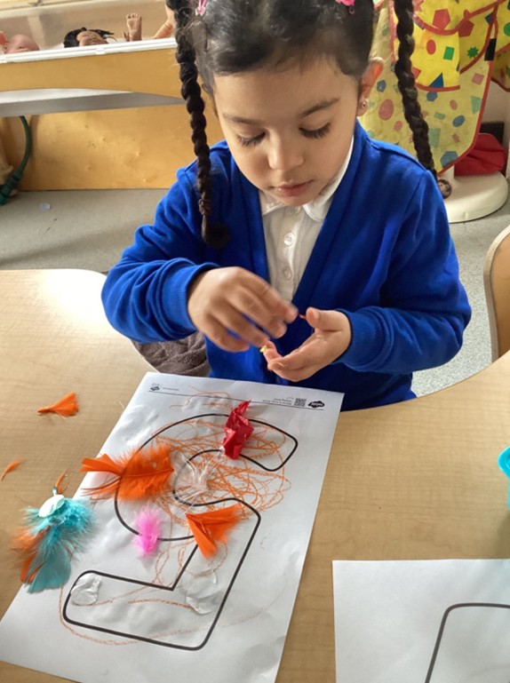 A girl with long dark braids concentrates as she glues colorful feathers and orange crayon scribbles onto a large printed number "5."