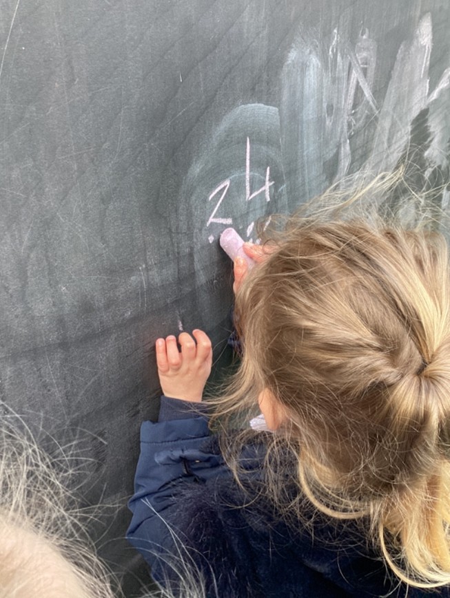 A view from behind a young girl with blonde hair as she uses a thick piece of purple chalk to write numbers on a large outdoor chalkboard.