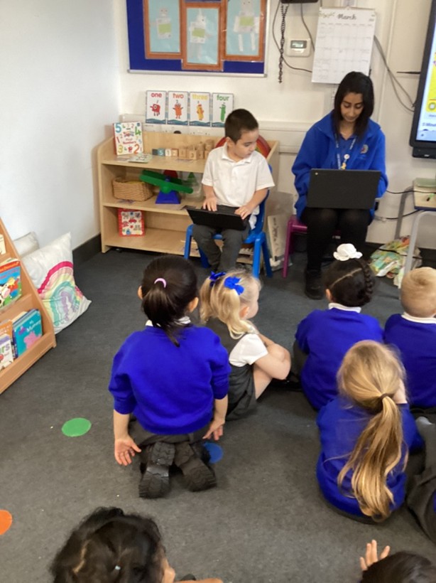 A teacher and a student sitting on chairs at the front of a room, both using laptops, while a group of students sits on the floor watching them.