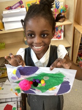 A smiling student holding up a craft project decorated with tissue paper and a cluster of colorful pom-poms.