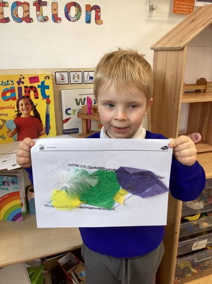 A student holding up a craft project decorated with green, purple, and yellow tissue paper, along with feathers.