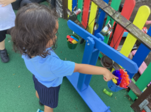 A child in a blue polo shirt points into a small blue bucket filled with bright orange and purple interlocking toys hanging from a blue wooden balance scale.