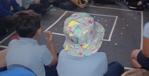 A child wearing a bucket hat covered in a colourful pattern of shapes sits on the carpet with classmates.